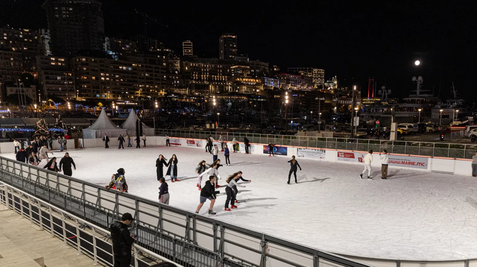Skaters spread across the open-air ice rink at Port Hercule at night, with the lit apartment blocks of Monaco rising behind the rail. — Patinoire du Port Hercule (Stade Nautique Rainier III), Monaco