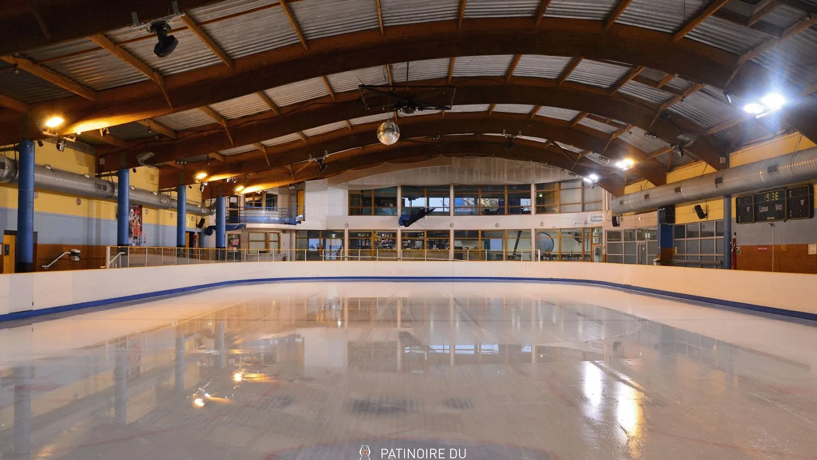 The empty ice from the boards, with an arched timber roof, ceiling spotlights and a mirror ball, and the glazed first-floor cafeteria along the far side. — Patinoire du Scorff