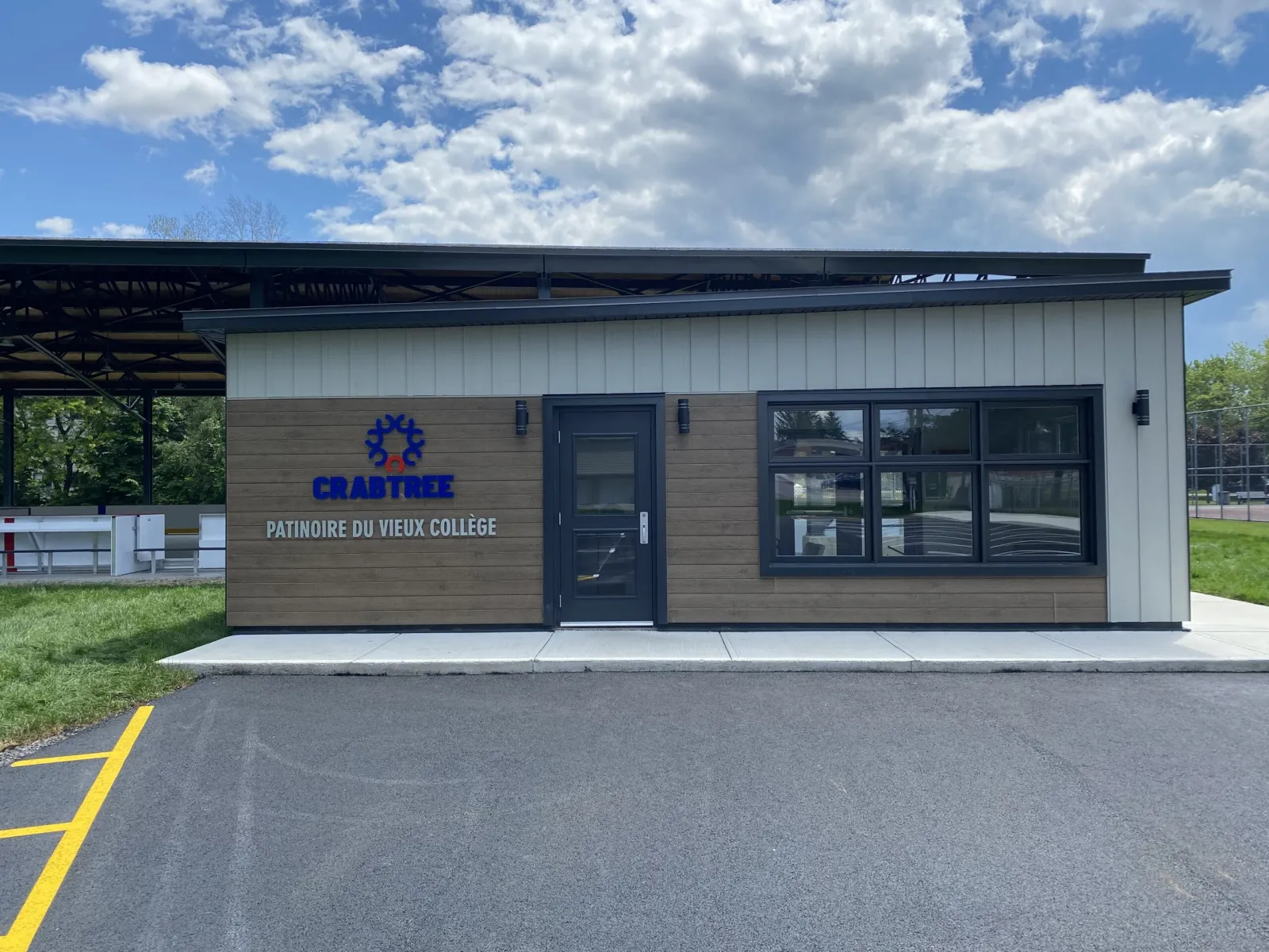 Le bâtiment de services de la Patinoire du Vieux Collège avec son enseigne, sous le toit couvert de la patinoire et à côté d'un stationnement asphalté. — Patinoire du Vieux Collège, parc Denis-Laporte (Crabtree)
