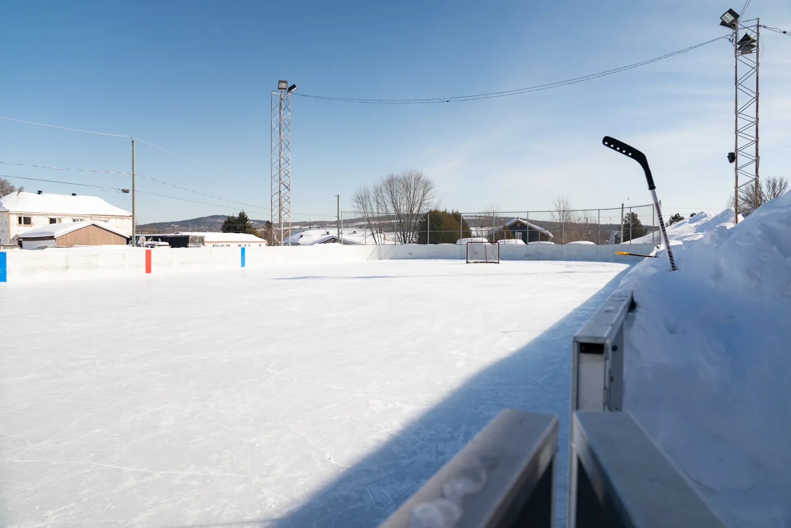 The empty boarded ice of the village rink in winter, with a goal at the far end, snowbanks along the boards and floodlight masts behind. — Patinoire du village (Saint-Ferréol-les-Neiges)
