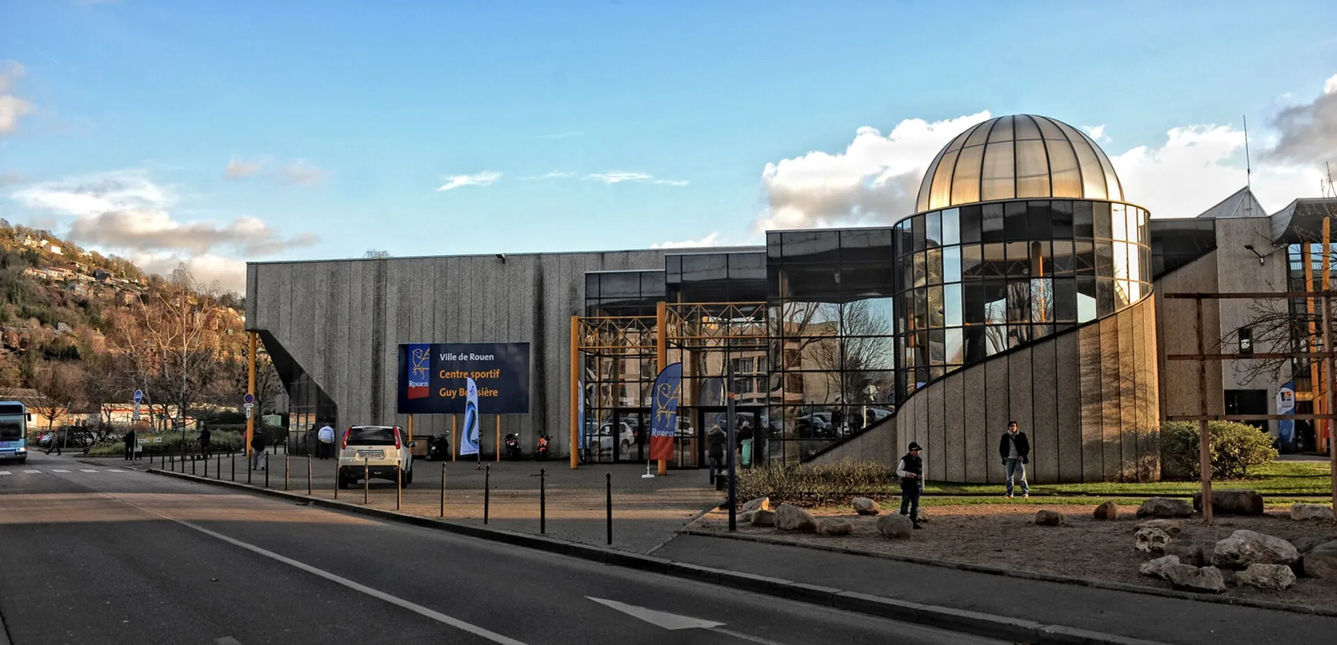 The street front of the Guy Boissière sports centre on the Île Lacroix in Rouen, which houses the city's ice rinks, with its concrete facade and glazed domed entrance. — Patinoire de loisirs Édith Ballester
