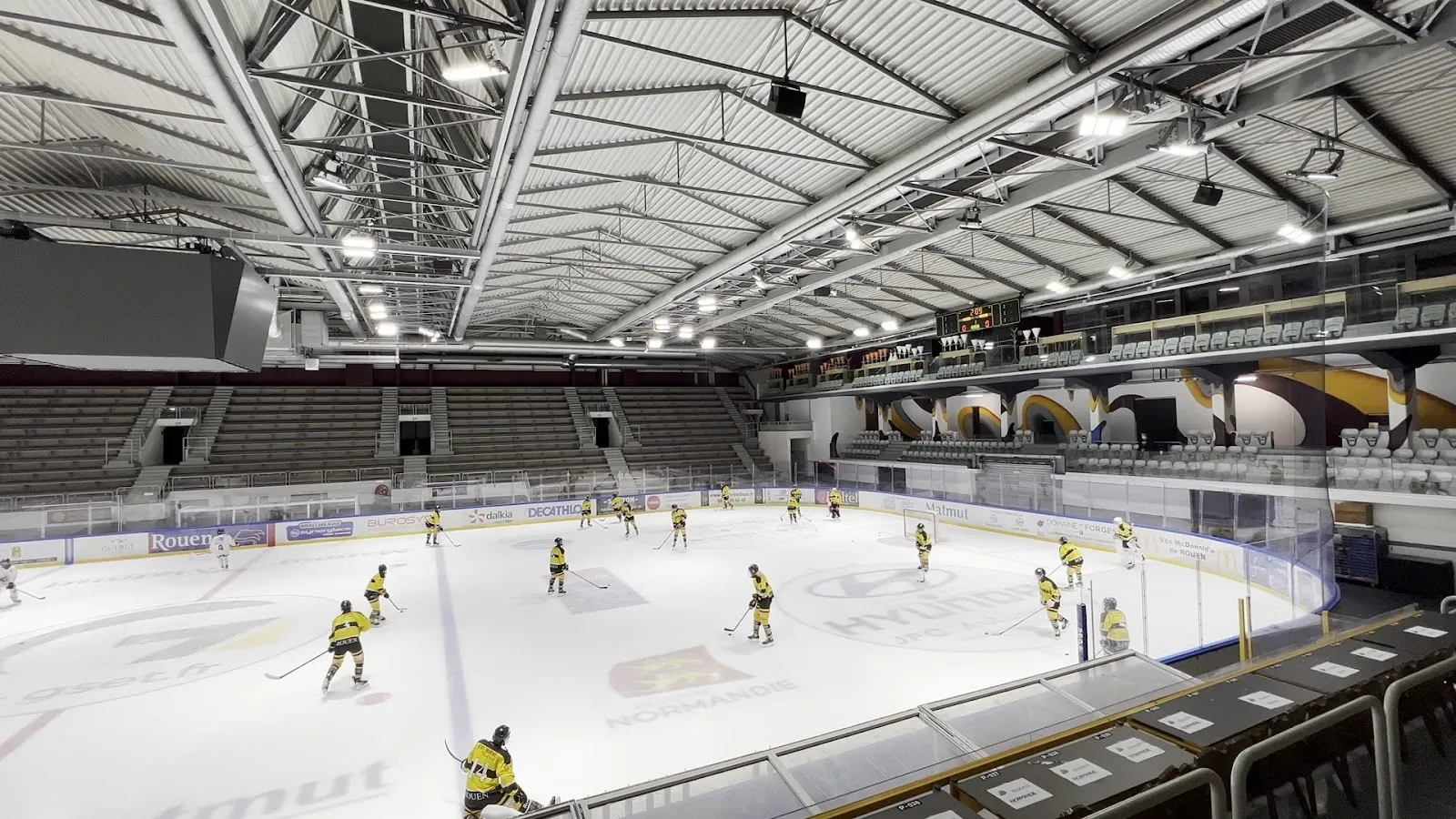 An ice hockey match on the Rouen rink seen from the stands, players spread across the ice under the steel-trussed roof with spectators along the far side. — Patinoire de loisirs Édith Ballester