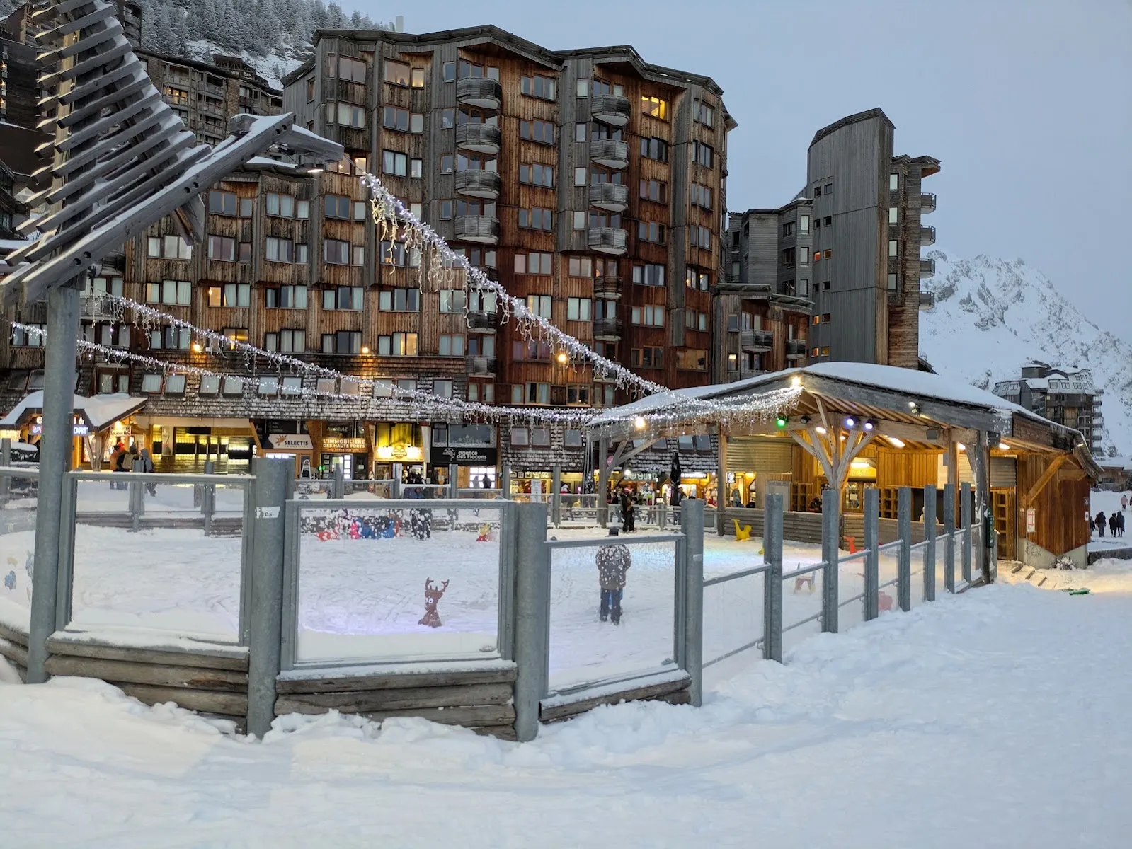 The fenced outdoor rink on the snow-covered central square at dusk, with the timber chalet-style kiosk and the resort's apartment blocks behind. — Patinoire en plein air d'Avoriaz