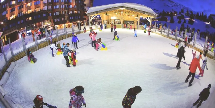 The outdoor rink in the central square at Avoriaz with skaters on the ice and the resort buildings and snow around it. — Patinoire en plein air d'Avoriaz
