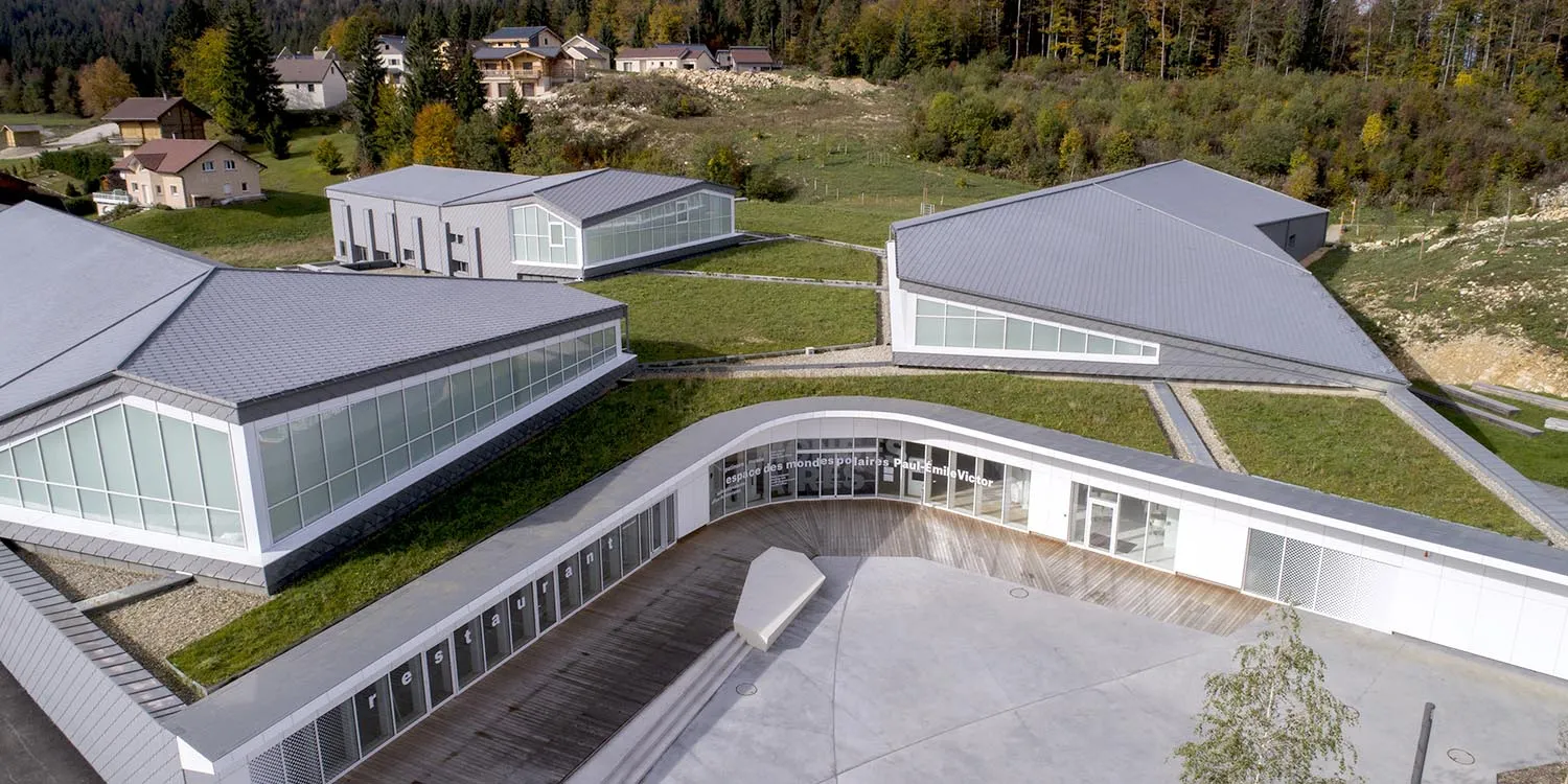 Aerial view of the Espace des mondes polaires in Prémanon: low grey-roofed pavilions with glazed fronts and green roofs, set on the Haut-Jura hillside — Patinoire de l'Espace des Mondes Polaires