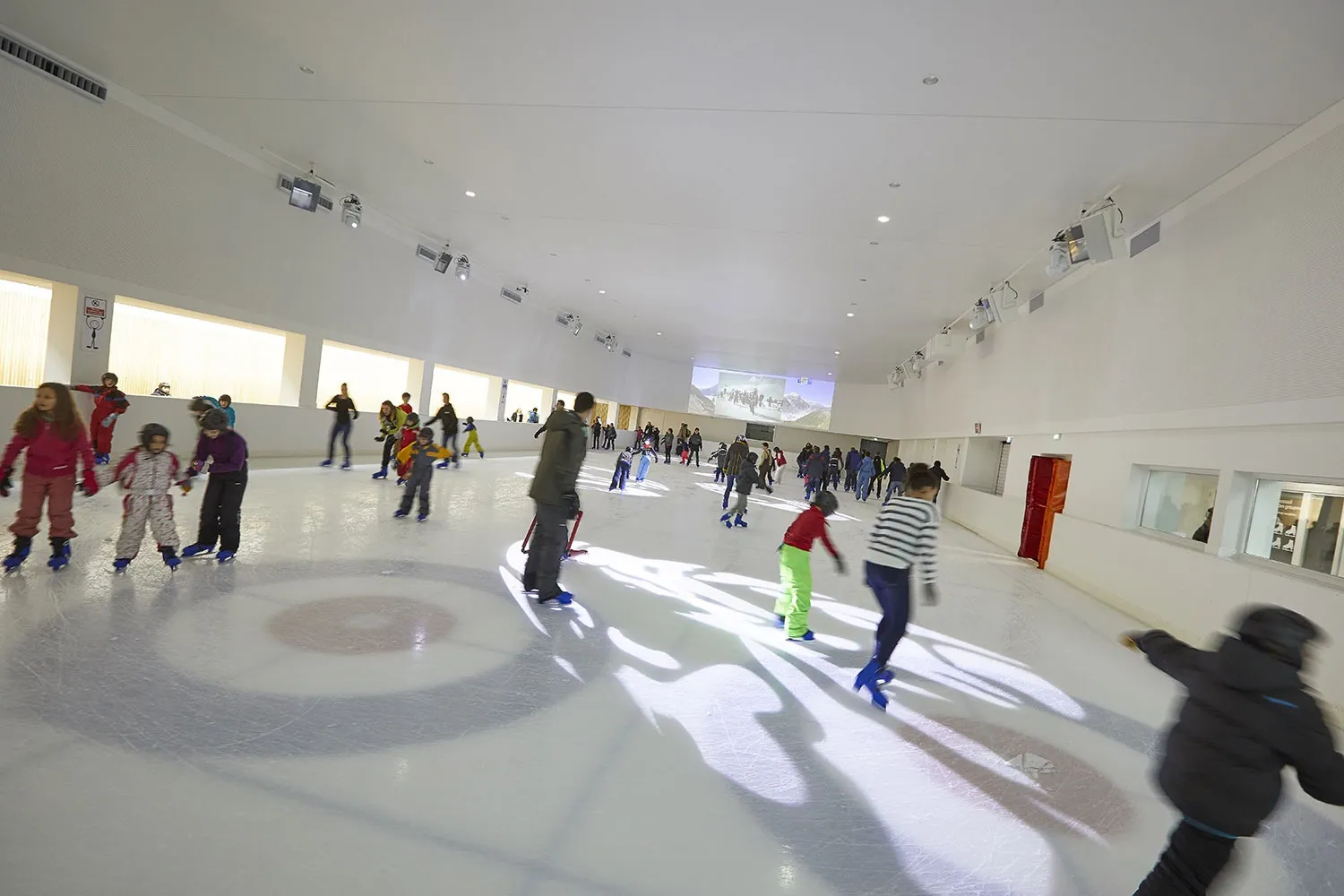 Children and adults skating across the pale rink at the Espace des Mondes Polaires, with a painted ring on the ice and the long hall behind. — Patinoire de l'Espace des Mondes Polaires