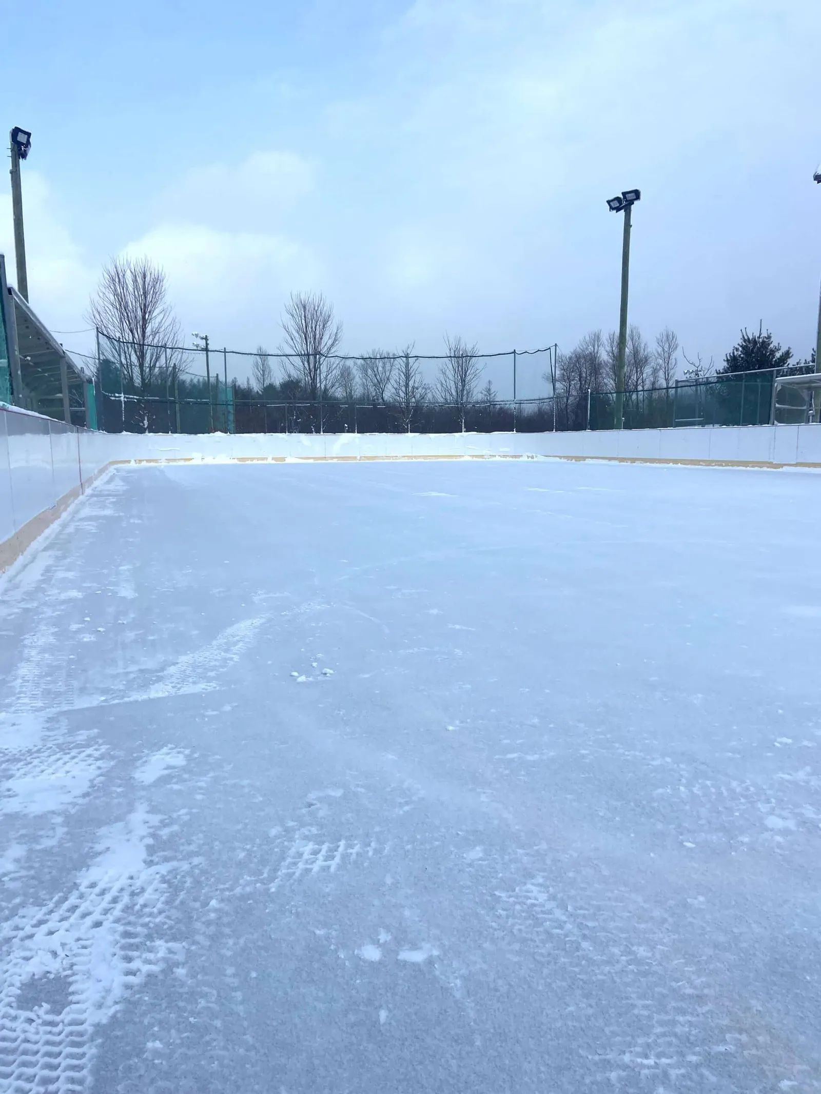 The Parc Goyette-Hill outdoor rink in Sutton on a sunny winter day, with the boarded ice sheet in the foreground and floodlight poles and bare trees beyond. — Patinoire et sentier de glace du parc Goyette-Hill