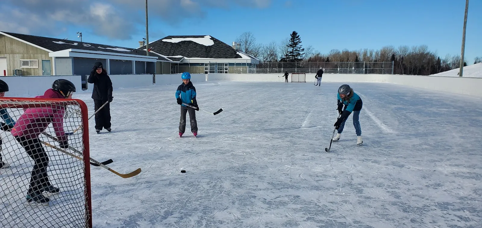 Three players in a pick-up game on the boarded outdoor rink at Pointe-au-Père with the park pavilion behind. — Patinoire extérieure de Pointe-au-Père