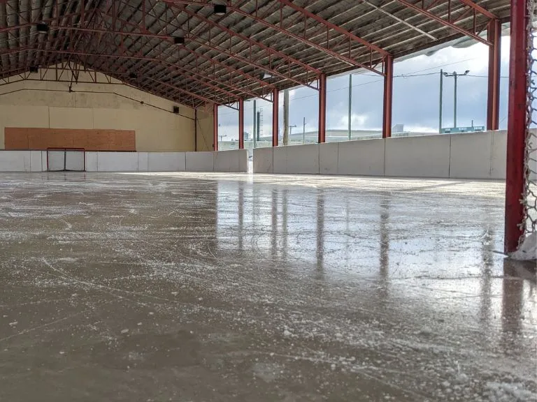 The covered outdoor rink in Saint-Agapit: a sheet of scuffed ice under a red steel-truss roof, with low white boards and open sides looking out over the village. — Patinoire extérieure de Saint-Agapit