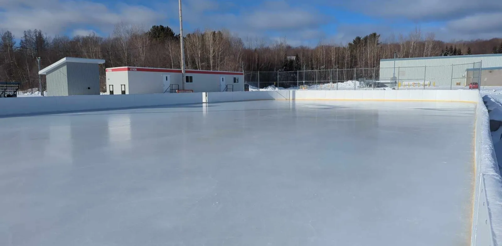 The empty boarded outdoor rink with clean ice, small buildings and bare trees behind the boards. — Patinoire extérieure de Saint-Boniface