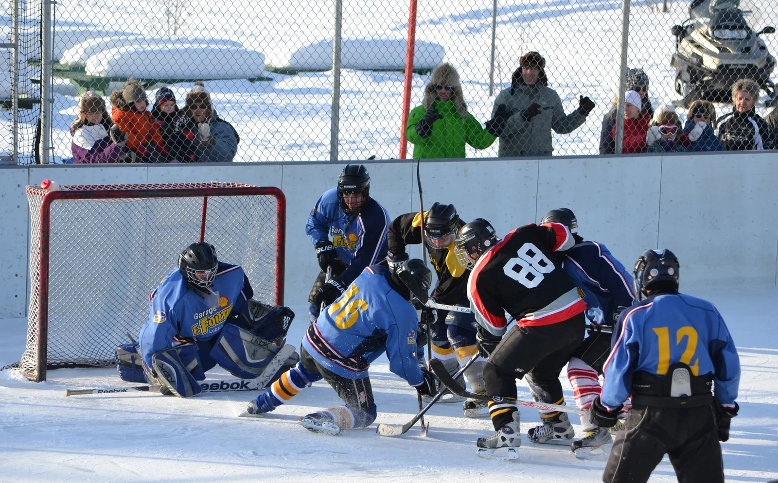 A hockey game on the outdoor rink, with players around the net and onlookers behind the boards. — Patinoire extérieure de Saint-Ludger-de-Milot
