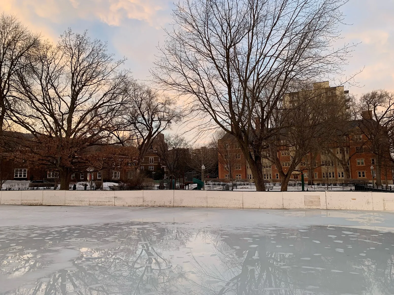 The boarded outdoor rink at Queen Elizabeth Gardens at dusk, with the playground and apartment blocks behind — Patinoire extérieure des jardins Queen Elizabeth