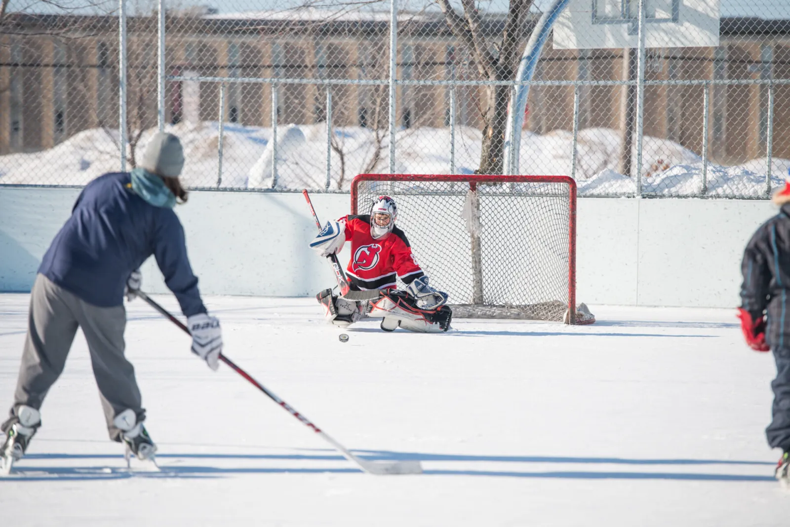A goalie and a shooter on the outdoor rink at the Parc récréatif in Montmagny, with the net, the boards and a fence behind. — Patinoire extérieure du parc récréatif (Montmagny)