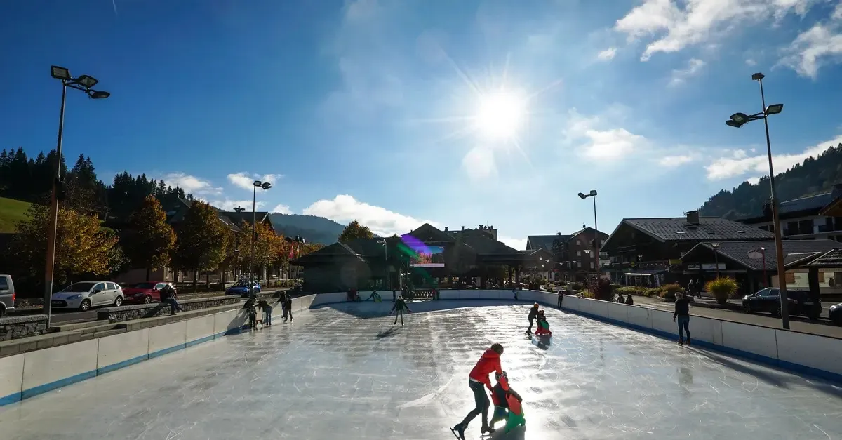 The open-air Les Gets rink on a sunny day, a few skaters on the ice with the village chalets and floodlight masts behind. — Patinoire extérieure des Gets