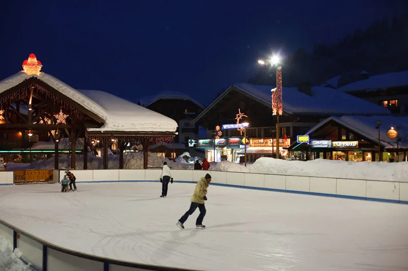 The outdoor rink at night in the village square, its white ice bordered by a blue line and the chalet buildings behind; skaters small in the distance. — Patinoire extérieure des Gets