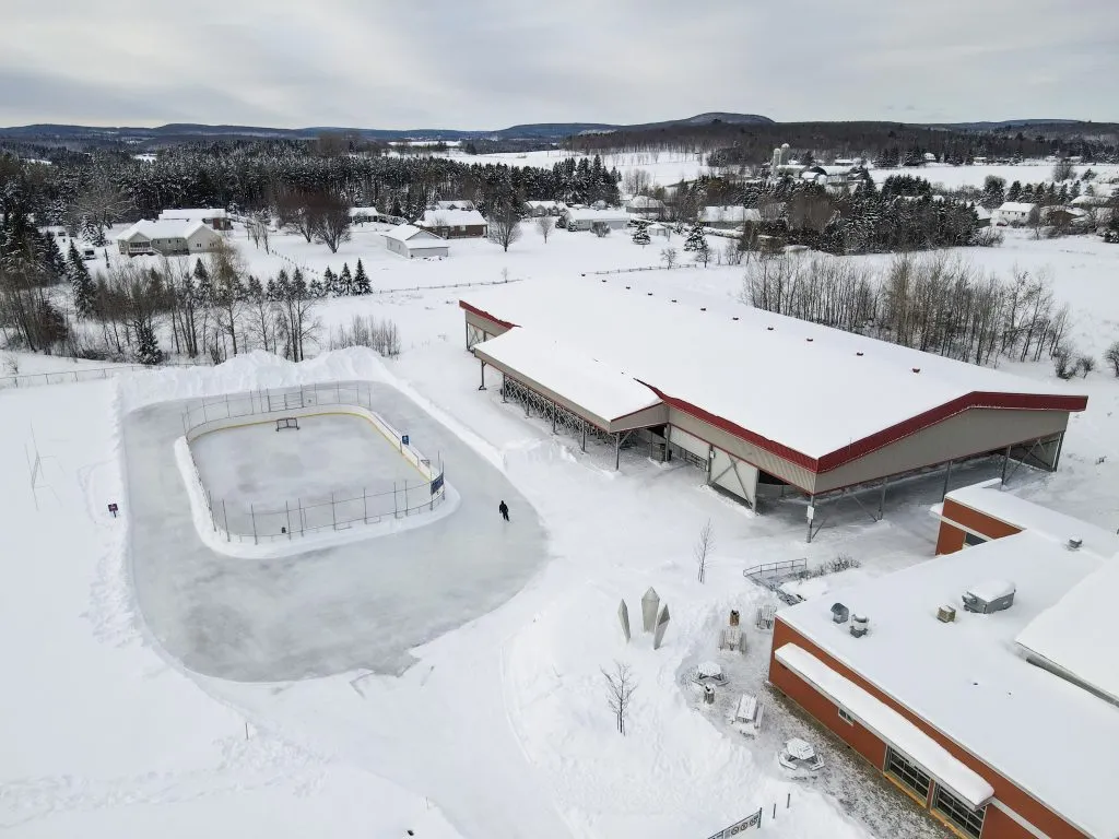 Aerial view of the open-air rink at Parc Donat-Gadoury in Saint-Jean-de-Matha in snow, beside the red-roofed arena buildings. — Patinoire extérieure non couverte du parc Donat-Gadoury (Saint-Jean-de-Matha)
