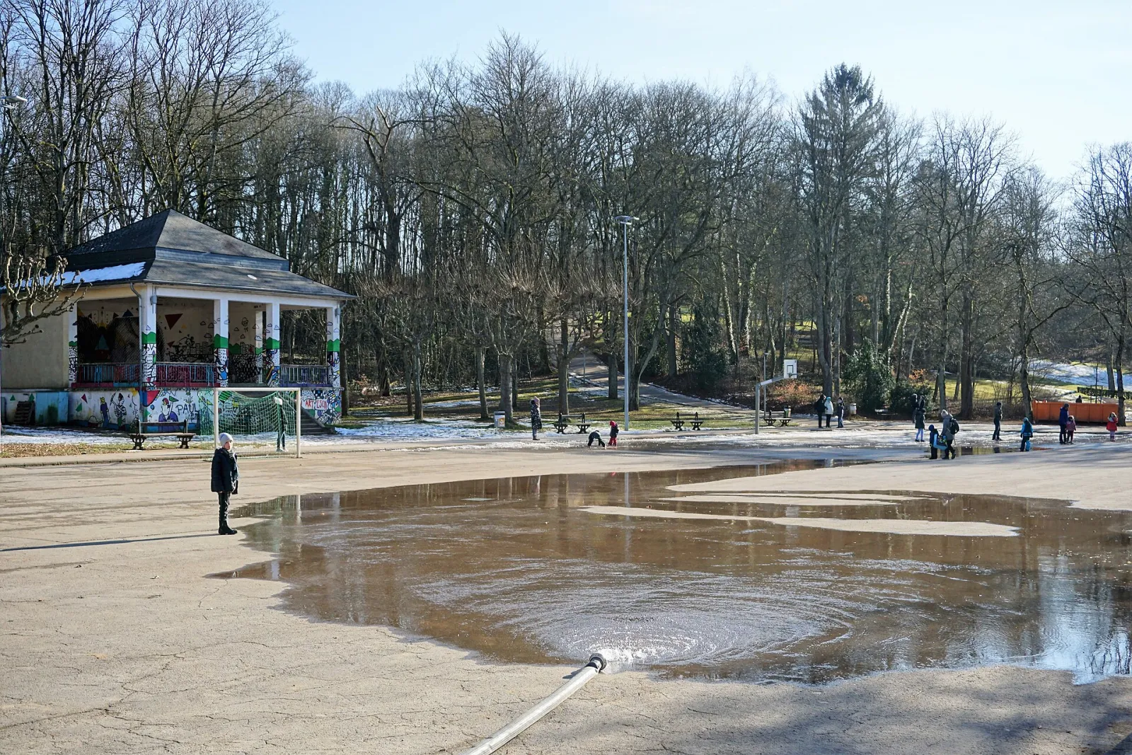 La zone de la patinoire Gaalgebierg en cours d’arrosage dans le parc municipal, avec le pavillon du kiosque à musique et le versant boisé derrière — Patinoire Gaalgebierg