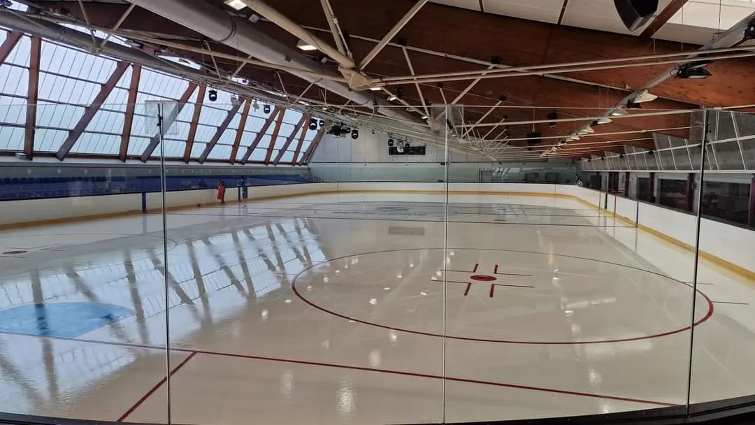 La glace vide vue à travers la vitre au bout de la patinoire, avec le rond de mise au jeu central, un logo bleu sur la glace et la toiture en bois et en acier avec son mur latéral vitré. — Patinoire de Mantes-la-Jolie