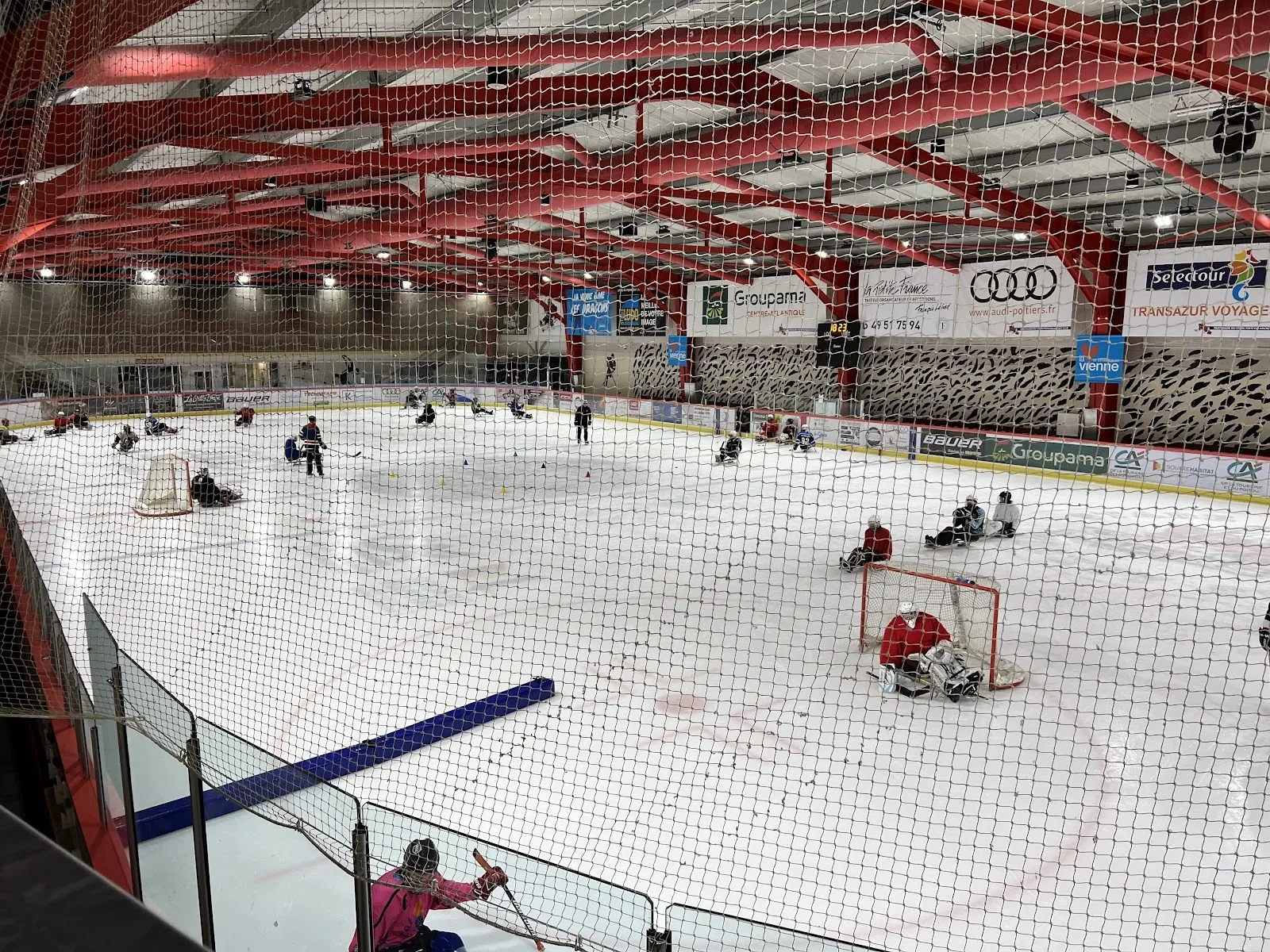 Vue large de la patinoire de Poitiers depuis les gradins : la glace avec des patineurs et un but, la charpente rouge et les panneaux de sponsors. — Patinoire de Grand Poitiers
