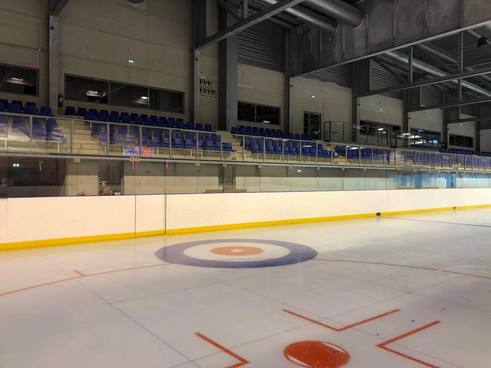 The empty ice sheet of the Halle Olympique with a curling target painted near the boards and the blue seating stands behind. — Patinoire de la Halle Olympique