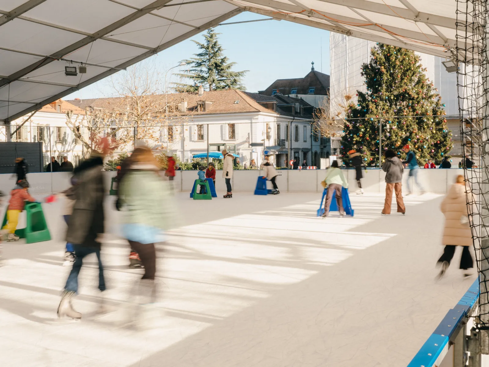 Skaters in motion on the white ice of the covered Carouge winter rink, with children using blue and green skating aids along the boards, a decorated Christmas tree and old town houses beyond the open side. — Patinoire hivernale de Carouge