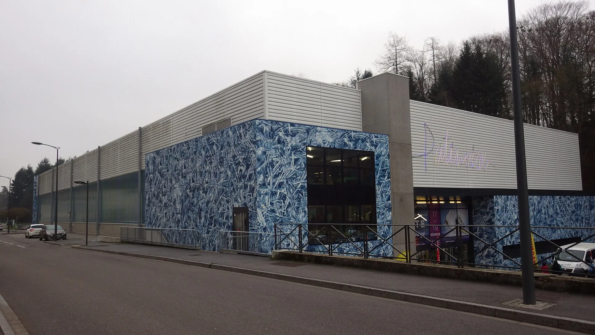 Le bâtiment de la patinoire d’Épinal vu depuis la rue, avec sa façade imprimée d’un bleu glacé, son bardage gris et le lettrage lilas Patinoire au-dessus de l’entrée. — Patinoire intercommunale d'Épinal