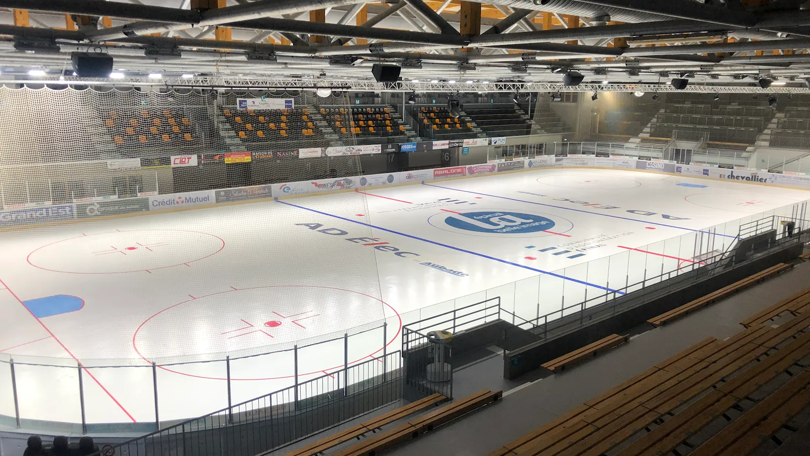 La glace vide et les gradins en bois étagés vus du haut de la tribune, avec les fermes de la toiture en bois et acier et les panneaux de sponsors. — Patinoire intercommunale d'Épinal