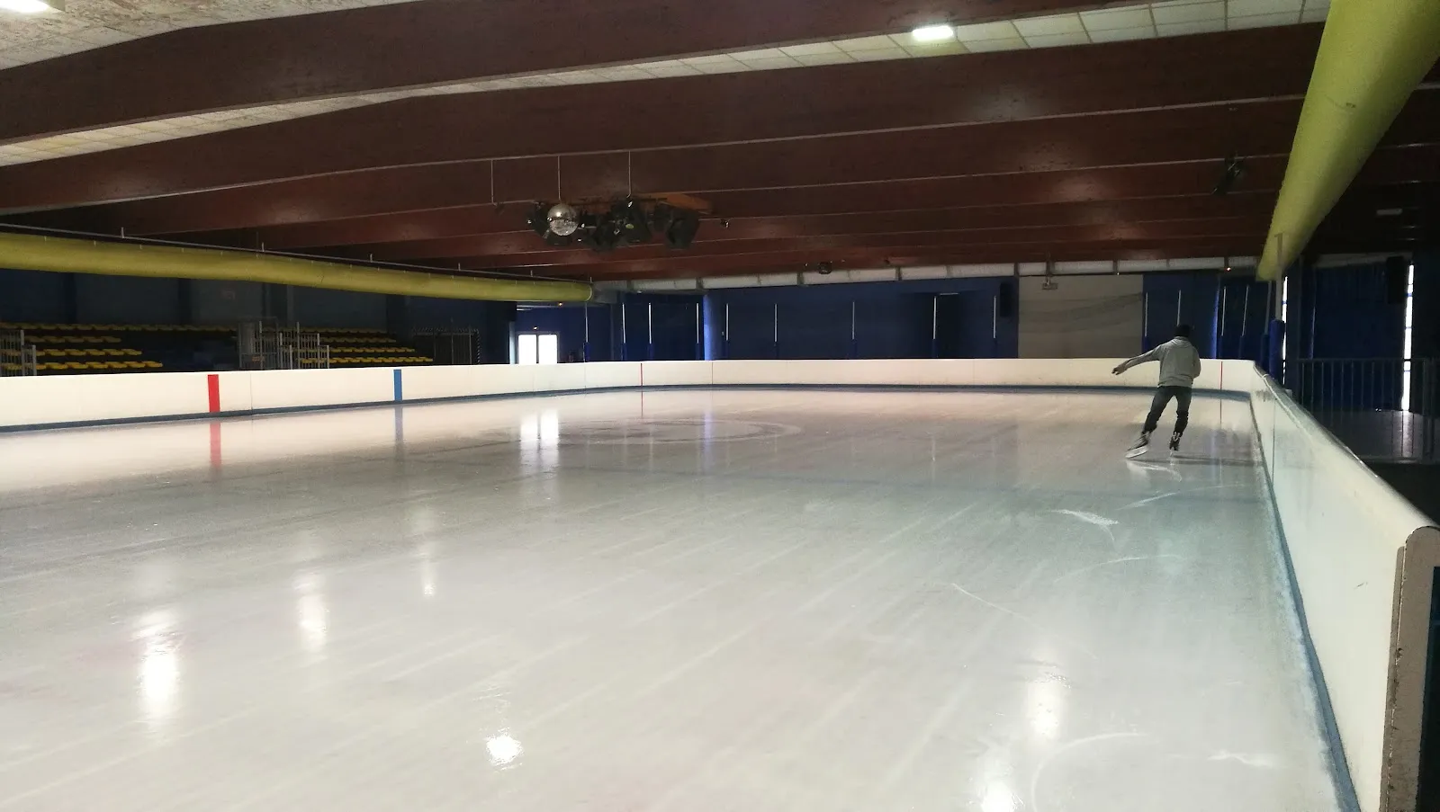 The almost empty ice sheet under a dark timber roof with a yellow air duct, a single skater at the far boards. — Patinoire d'Issoudun