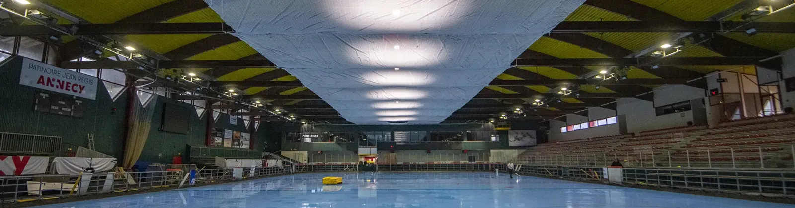 La glace et la charpente en bois de la patinoire Jean Régis, vues depuis le bout de la piste pendant les travaux de rénovation, avec la banderole « Patinoire Jean Régis Annecy ». — Patinoire Jean Régis