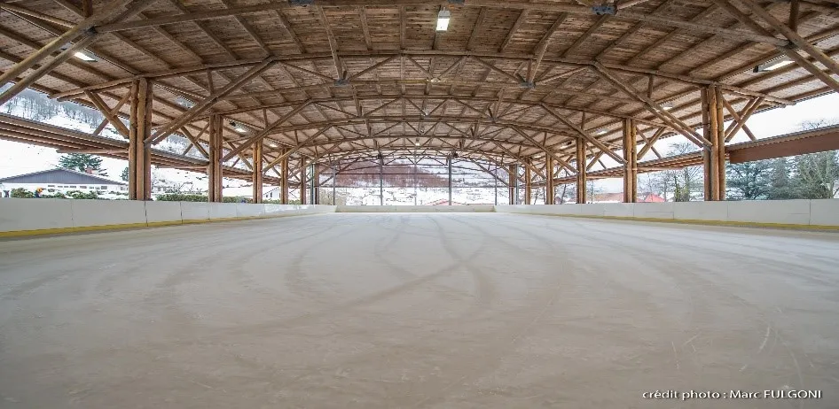 The empty ice sheet under the long open timber hall, with the wooden roof trusses, the low boards and the hillside beyond — Patinoire de La Bresse