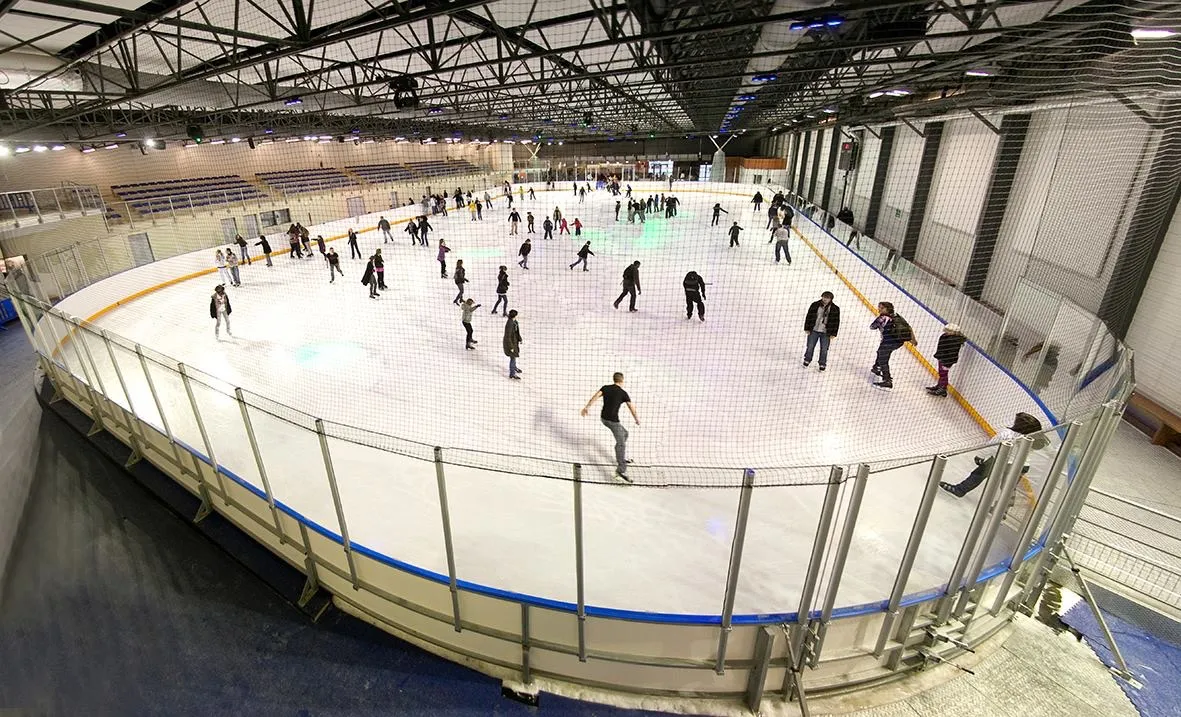 A wide view along the oval ice sheet from a corner of the boards, with a dozen skaters spread across the ice under a steel-truss roof; the boards are white with blue and yellow trim. — Patinoire de la Cartonnerie