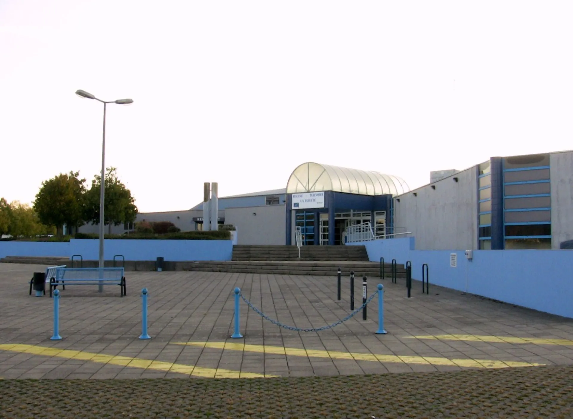 L’entrée du complexe piscine et patinoire Lafayette à Planoise, avec son porche vitré en dôme, le parvis pavé et les murs et potelets bleus. — Patinoire La Fayette
