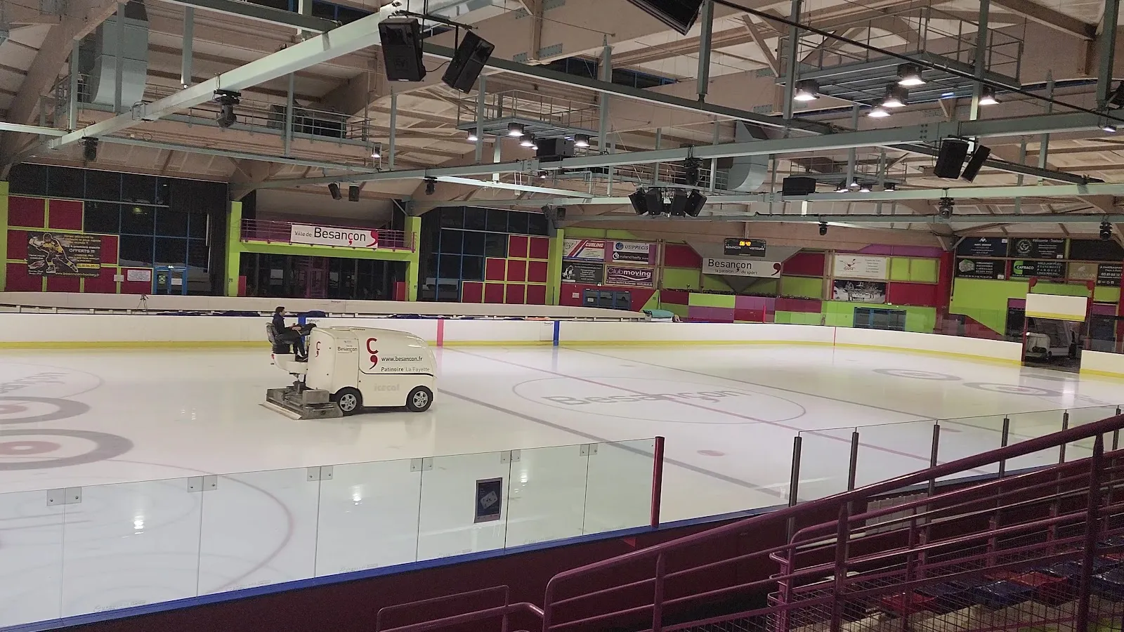 La grande patinoire vue des gradins, avec la surfaceuse à glace au travail sur la glace vide, des sièges rabattables rouges à droite et des panneaux verts et rouges sur le mur du fond. — Patinoire La Fayette