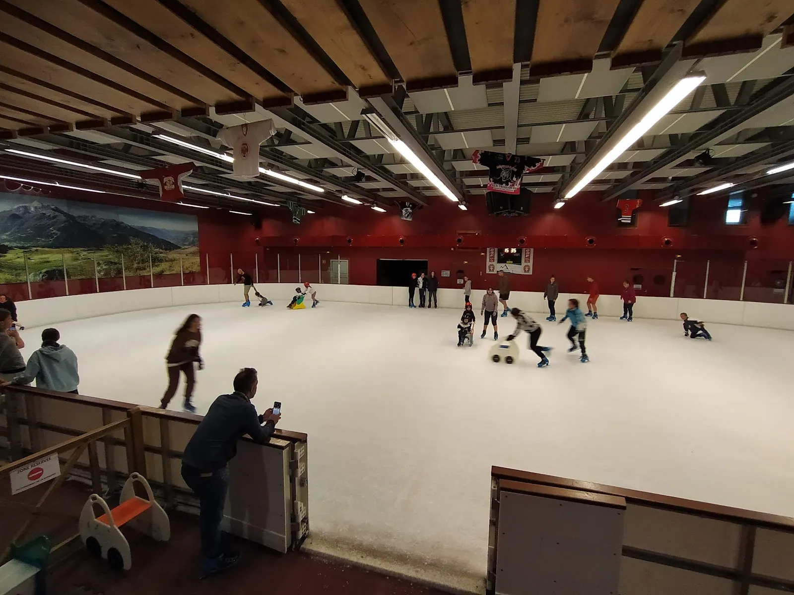 The covered ice with a timber ceiling and a red wall carrying a mountain mural, with a few skaters and children using skating aids across the rink. — Patinoire de La Rosière
