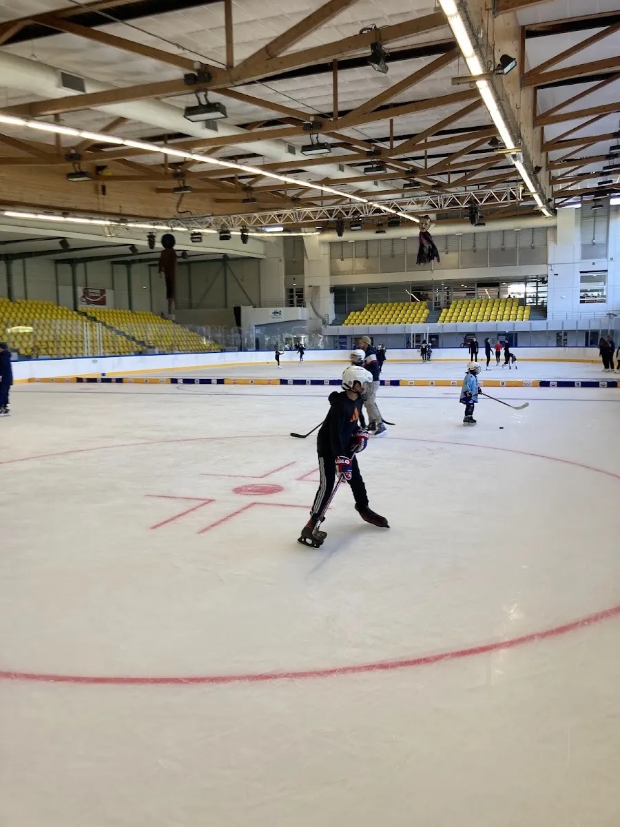 Des patineurs et de jeunes joueurs de hockey sur la glace de Castres Sports de Glace, avec les fermes en bois du toit au-dessus et des sièges jaunes le long du côté opposé. — Patinoire de l'Archipel