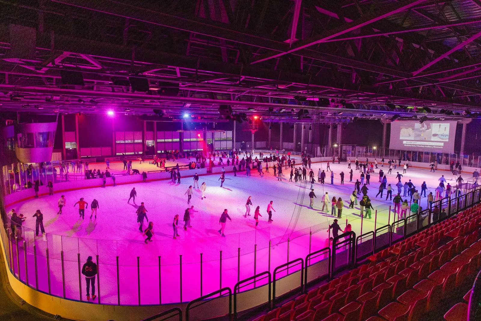 The Blizz's main rink lit in pink and purple during an evening session, with skaters on the ice and the red tip-up seats along the boards — Le Blizz