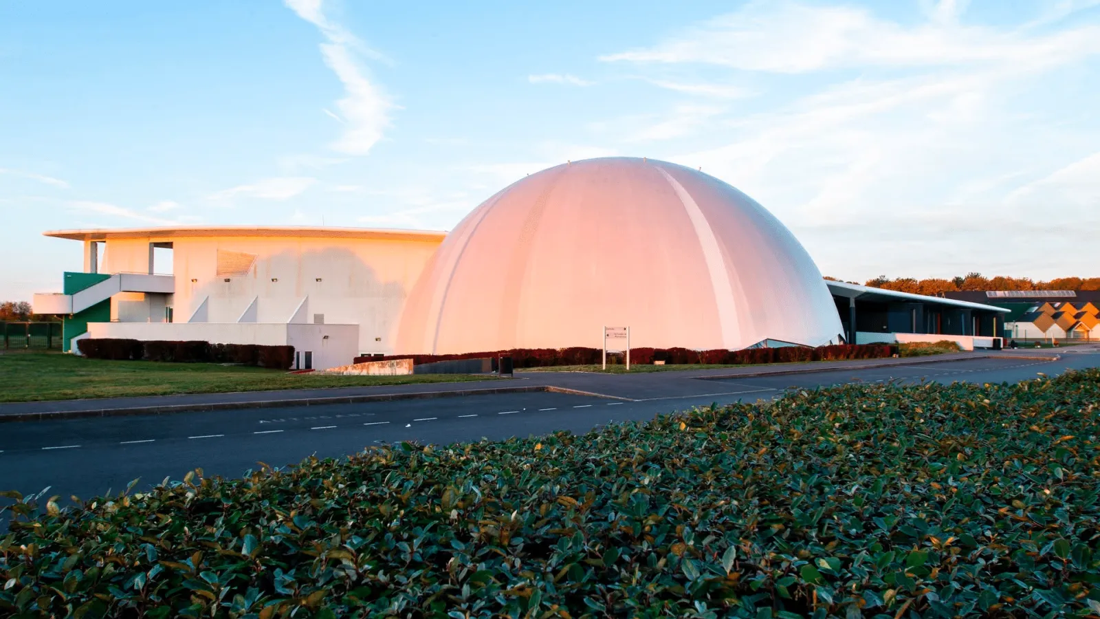 The white dome of the complex lit by low sun, with a flat-roofed white wing beside it, seen across the road and a hedge. — Patinoire du Dôme