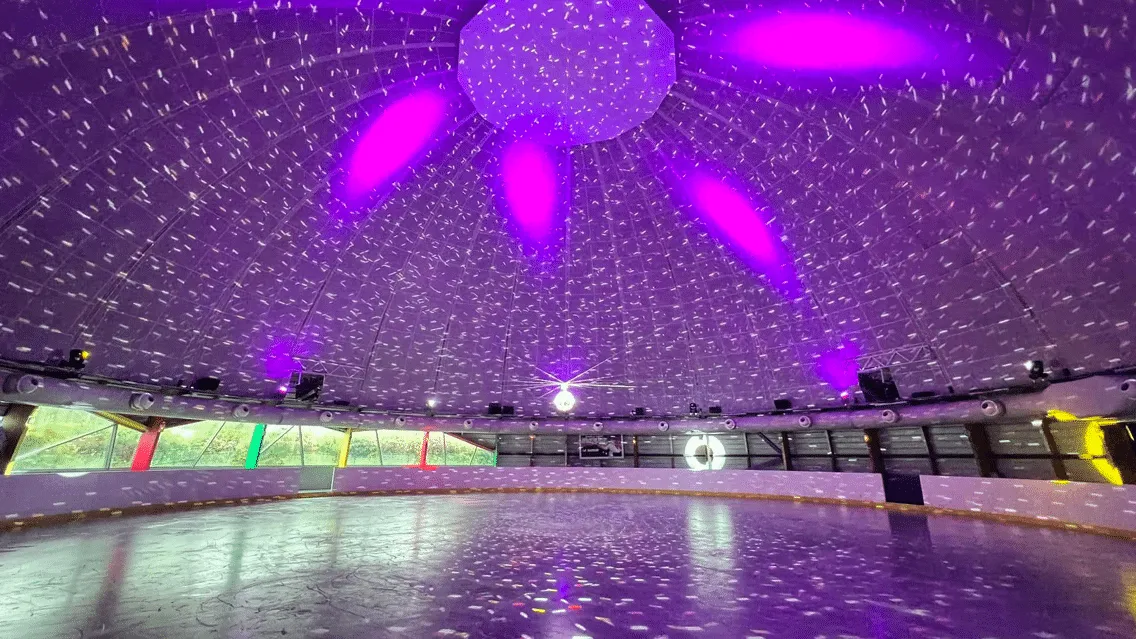 The empty ice seen from rinkside under the underside of the dome, with purple light effects scattered over the ceiling and the surface. — Patinoire du Dôme