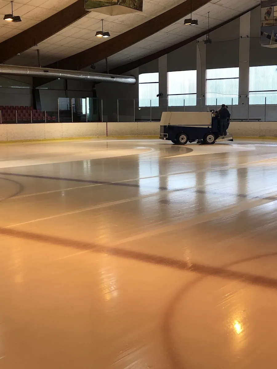 La glace de la patinoire du Havre vue depuis le bord de la piste, avec la surfaceuse à glace au travail contre les bandes du fond, sous les poutres de la toiture en bois. — Patinoire du Havre