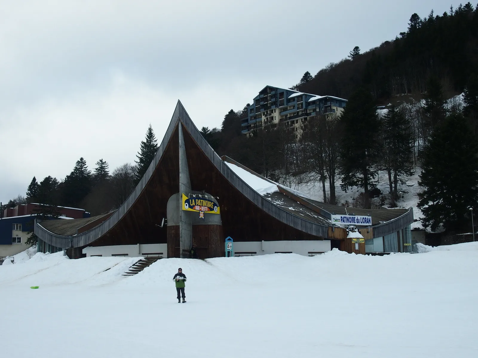 The curved wooden roof of the patinoire building rising above snow-covered ground, with its entrance sign and wooded slope behind. — Patinoire du Lioran