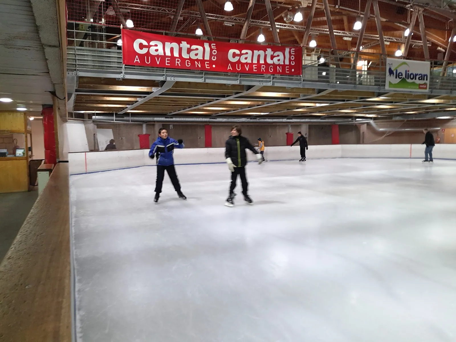 Skaters on the ice sheet of the Le Lioran rink, seen along the boards, with the timber-roofed hall and the Cantal Auvergne and Lioran banners above. — Patinoire du Lioran