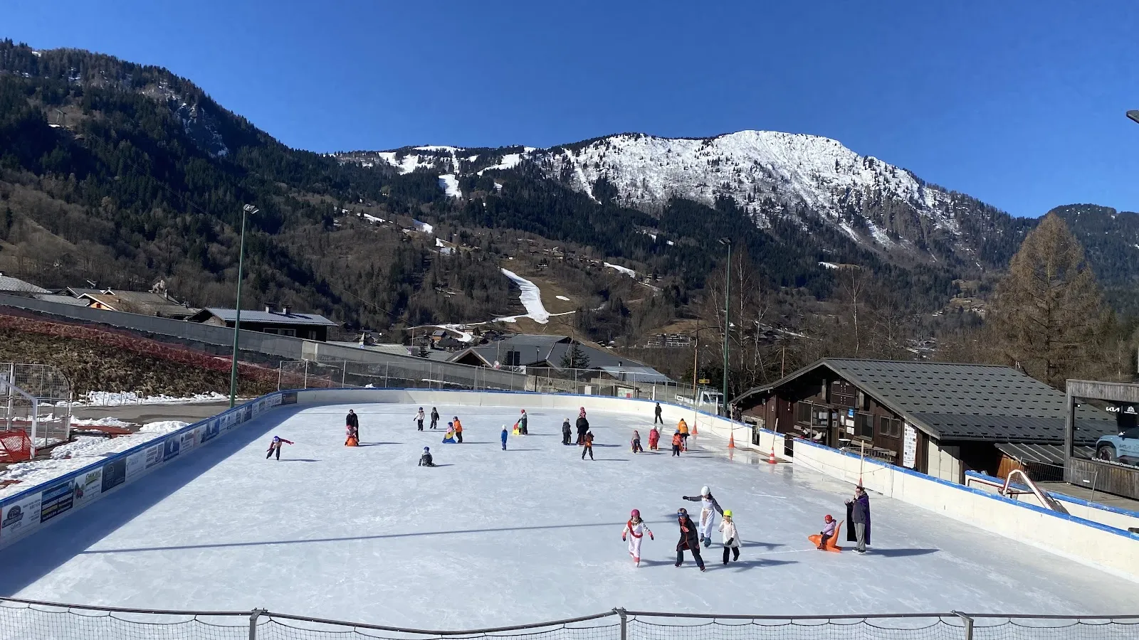 The open-air rink beside the Houches valley road, ringed by boards with a timber chalet building and the snow-streaked mountain slopes behind. — Patinoire des Houches