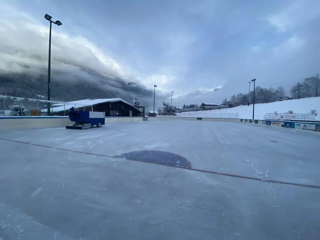 The Les Houches open-air ice with its resurfacer and floodlights, under cloud with the valley slopes behind. — Patinoire des Houches