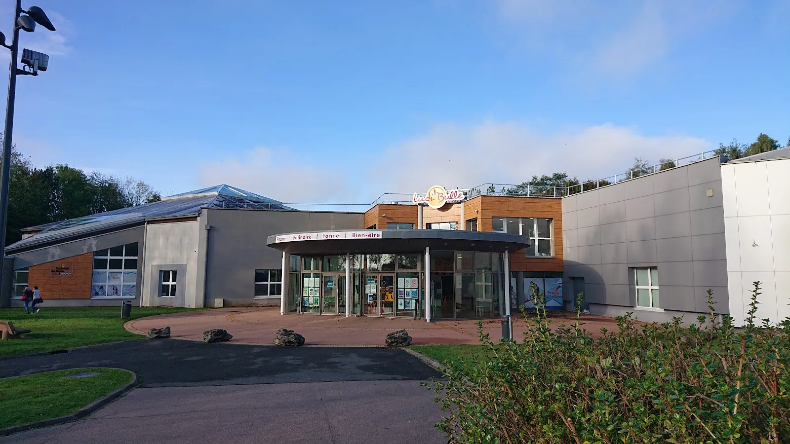 The low entrance building with its round glazed canopy and grey and timber cladding, seen across the forecourt under a blue sky. — Patinoire Ludi'Bulle