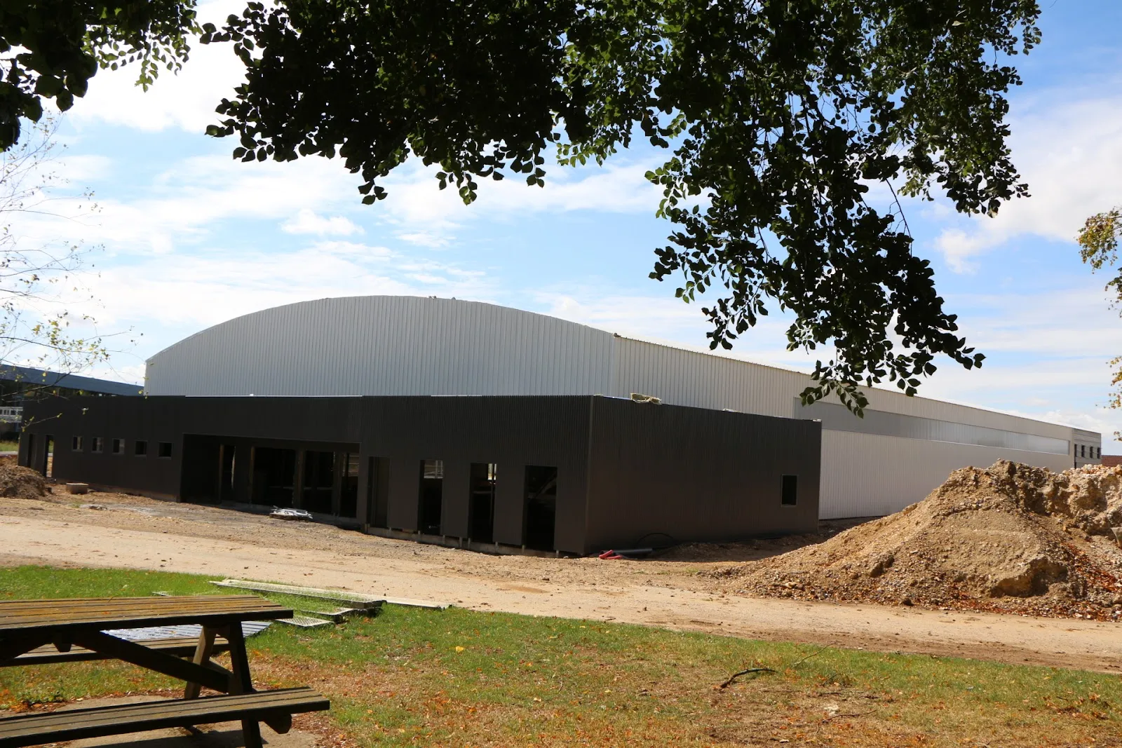 The rink building seen from the park, with its grey arched roof and dark cladding, a picnic table and bare earth in the foreground. — Patinoire Marcel Dassault