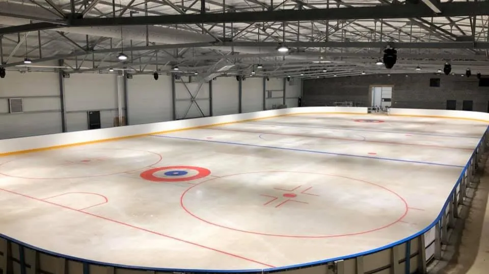 The empty ice from the rink-side, with the red circles and lines, the blue-and-yellow boards and the steel-framed roof of the hall. — Patinoire Marcel Dassault