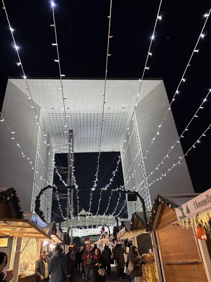 The Christmas market on the parvis of La Défense under strings of lights, with the Grande Arche behind the chalets — Patinoire du marché de Noël de Paris La Défense