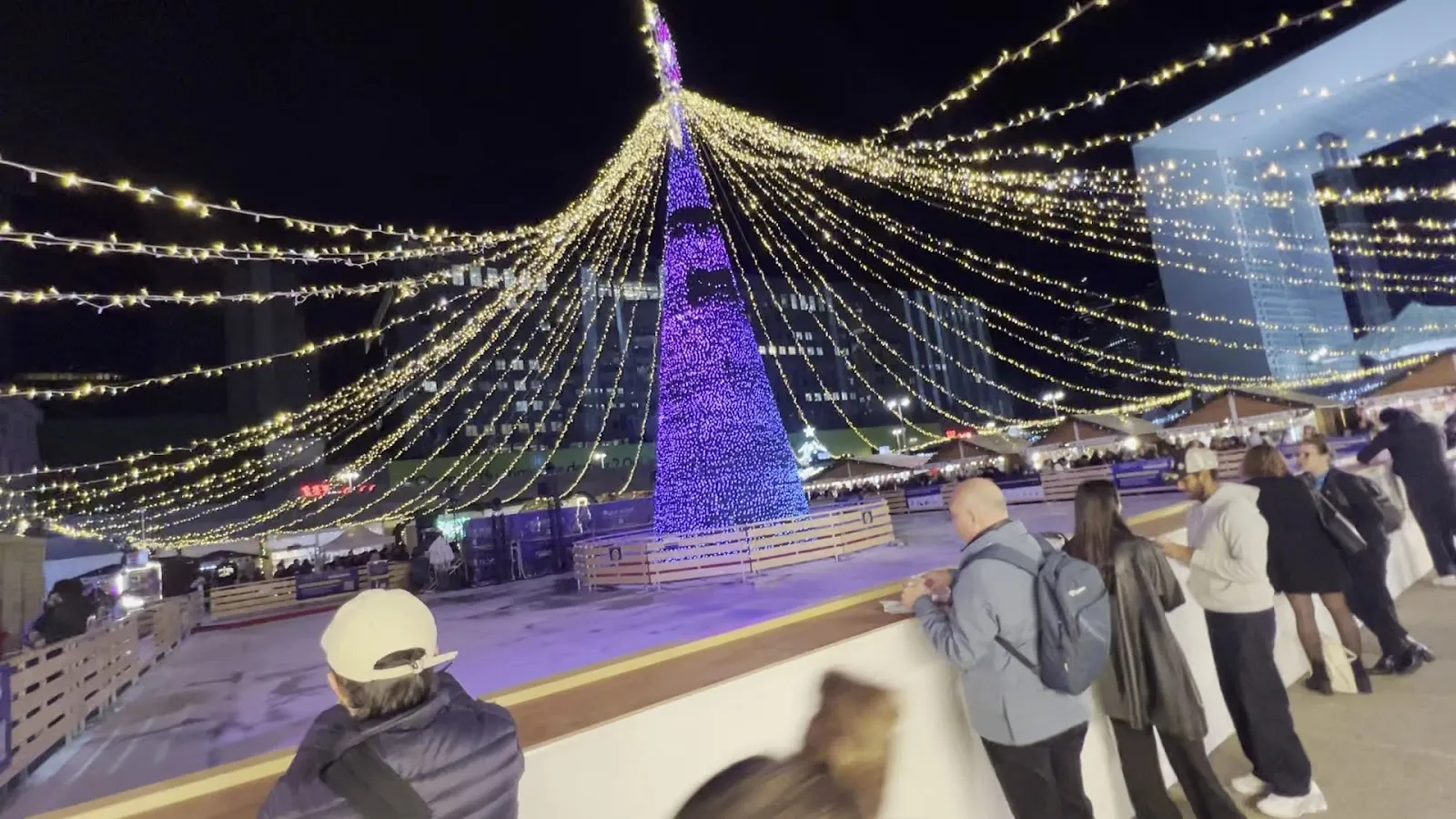 The corner of the La Défense Christmas-market rink, with people along the boards under the lit tree — Patinoire du marché de Noël de Paris La Défense