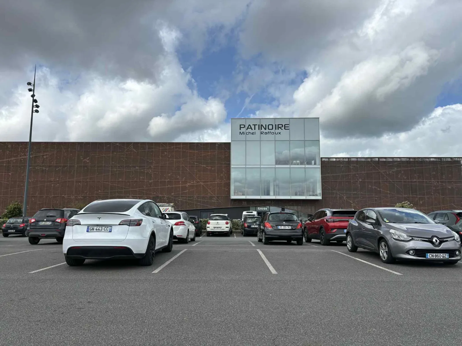La façade en métal brun du bâtiment, avec un bloc vitré portant l’enseigne PATINOIRE Michel Raffoux et l’entrée en dessous, vue depuis le parking sous un ciel nuageux. — Patinoire Michel Raffoux