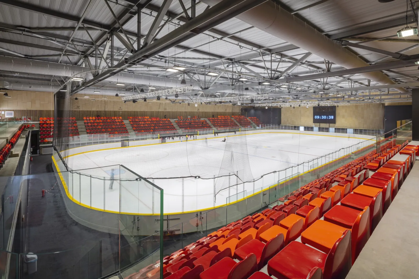 La piste de taille olympique vue des gradins : sièges rabattables rouges le long des bandes, un tableau d’affichage sur le mur du fond et une toiture à fermes d’acier, avec la glace vide. — Patinoire Michel Raffoux